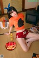 A woman in an orange shirt and red shorts sitting on the floor.